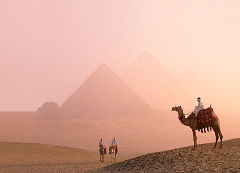 Three people riding camels with they pyramids of Giza, Egypt in the background