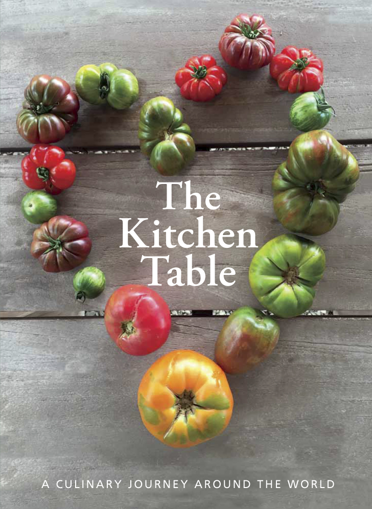 Photo of an image with tomatoes of all sizes and colors lined up in a shape of a heart and big texts that read "The Kitchen Table - A Culinary Journey Around the World"